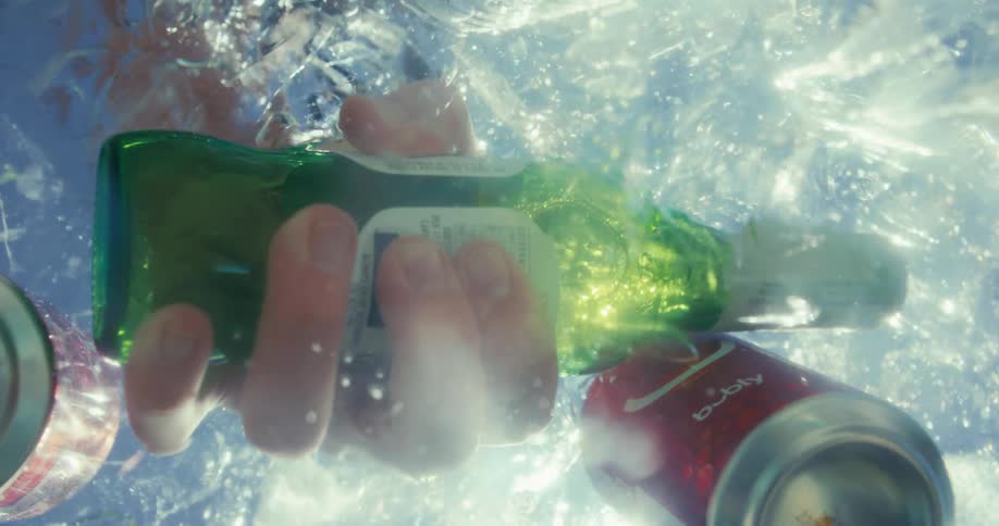 A persons arm reaching into icefilled water picking up a bottle of beer with two cans of drinks floating nearby in the water at midday