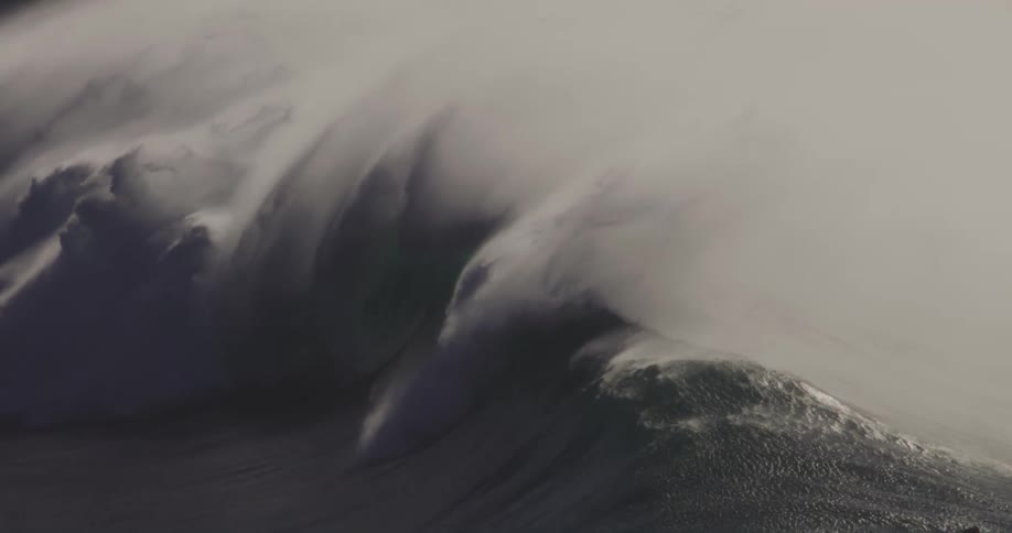 A massive dark green wave with white foam crests and barrels as it approaches a rocky shoreline in the background
