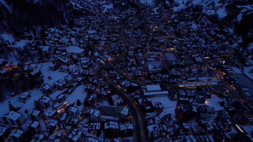 An aerial view of a snow covered town nestled in a valley between dark imposing mountains at dusk with scattered lights illuminating the buildings