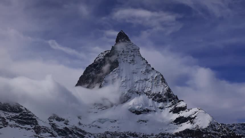 Clouds float by in a blue sky past one snowcovered pointy mountain
