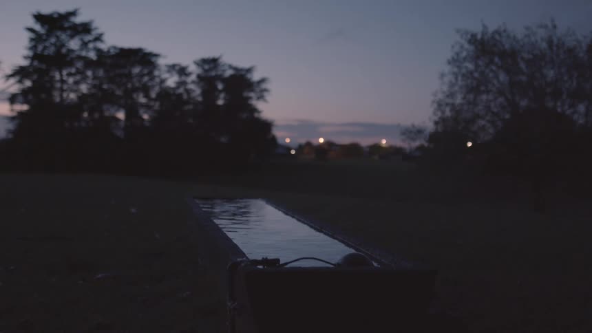 A narrow reflective body of water with silhouetted trees and distant lights against a dusky sky