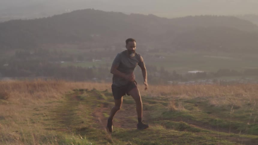 A man with a beard runs strenuously uphill on a dirt path in a dry grassy landscape with hazy mountains and a valley visible in the distant background under warm diffused light