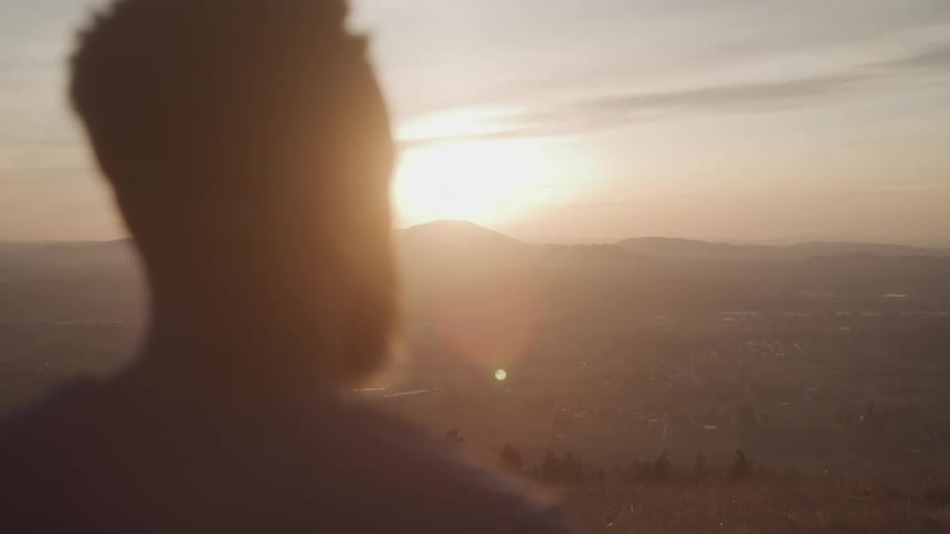 A silhouetted person is framed in the foreground looking out over a vast hazy valley and distant mountains bathed in the warm low light of a rising or setting sun