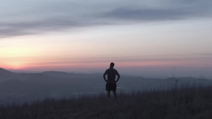 A man standing alone on a grassy hill in the evening as the sun sets behind him