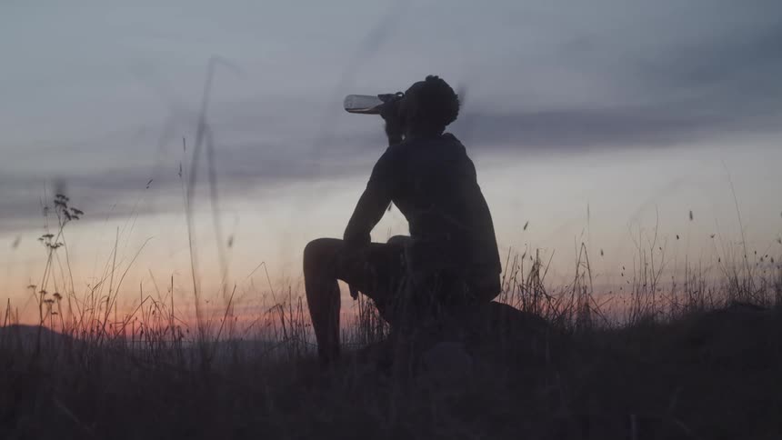 A man sitting on a log in a grassland drinking water from a bottle during the evening sunset