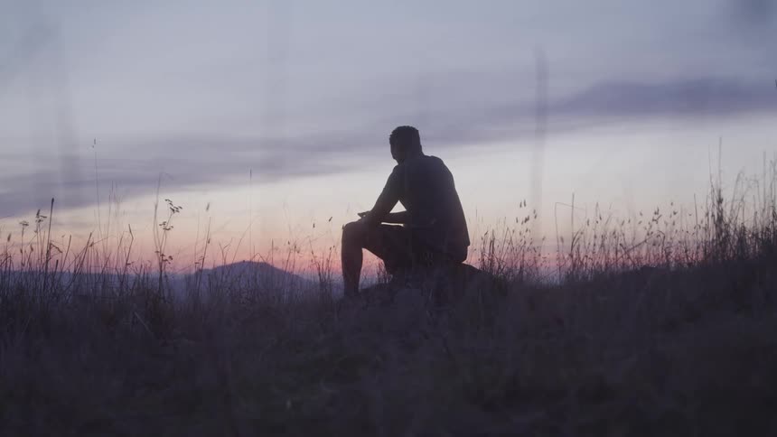 A man sits on a large rock in a field