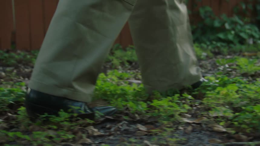 A man wearing black shoes and trousers walking on a lawn covered with grass at midday