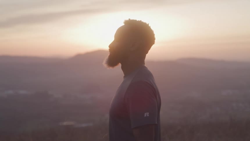 A person with a beard is standing outdoors at sunset with mountains visible in the background