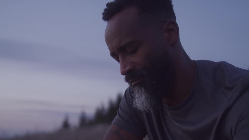 A black man sitting in a grassland at sunset with a mixed black and white beard and his eyes closed in a peaceful expression