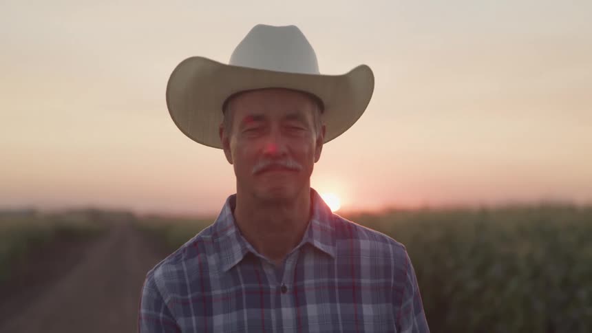 Older farmer smiles while standing near a field of crops at sunset