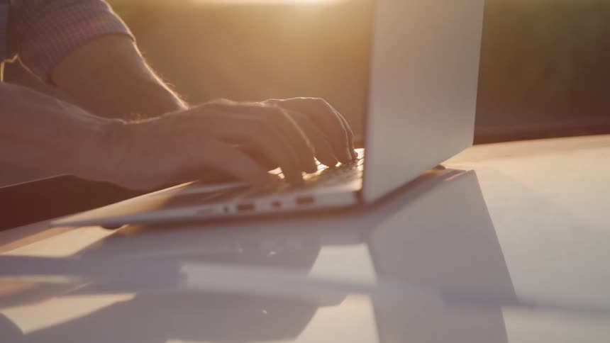 Close up of a man typing on a laptop computer on a vehicle hood outdoors at sunset