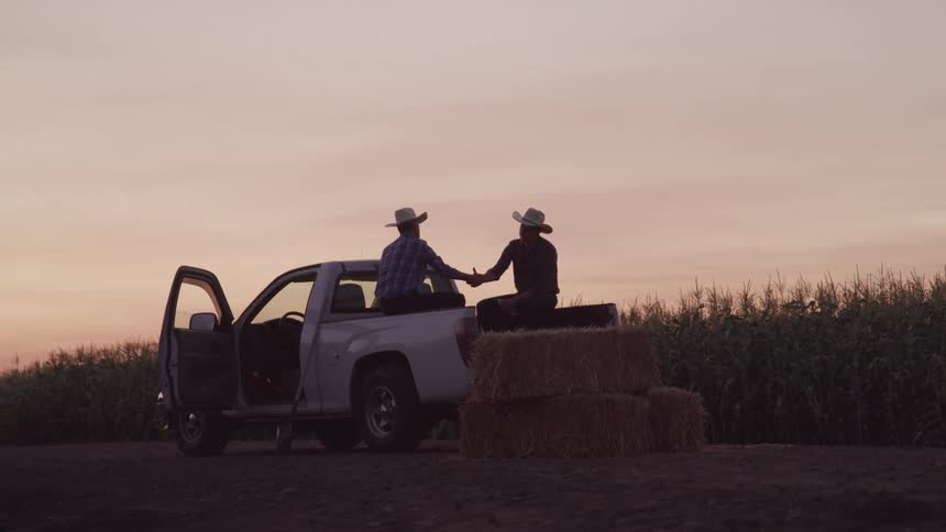 Men shake hands while sitting in the bed of a truck parked near a crop field under a red sky