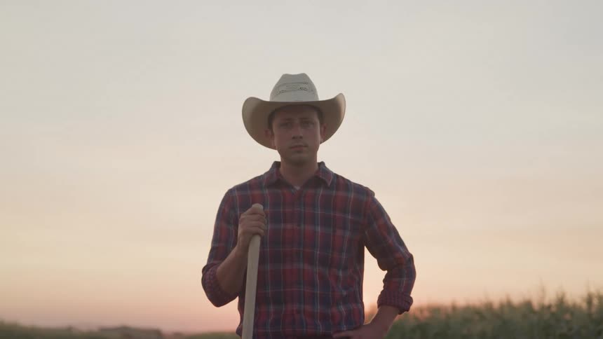 A farmer stands with his hand on his hip while holding something with a wooden handle in the other hand