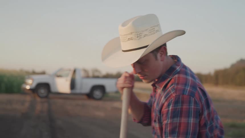 A man in a cowboy hat and plaid shirt bends down to work on a dirt road with a white pickup truck and bales of hay visible in the blurred background under warm low sunlight