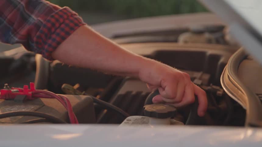 A hand in a red plaid shirt reaches into a vehicles engine bay inspecting its components