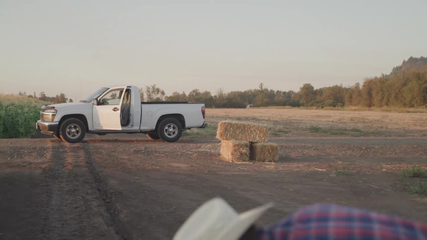 A farmer digging the soil under the bright midday sun with a truck parked in the distance and several bales of straw stacked beside it