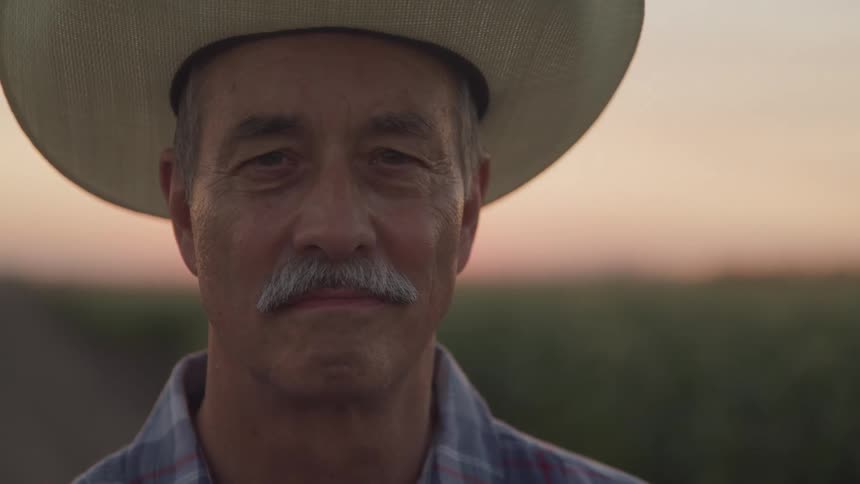 A middleaged man with a mustache wears a cowboy hat and a plaid shirt looking directly at the camera with a sunset field in the background