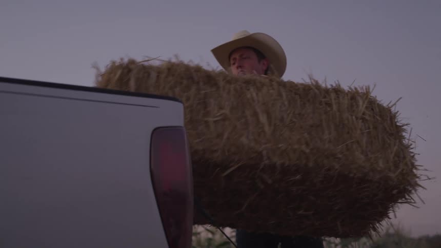 A man wearing a cowboy hat loads a hay bale into the back of a pickup truck at dusk