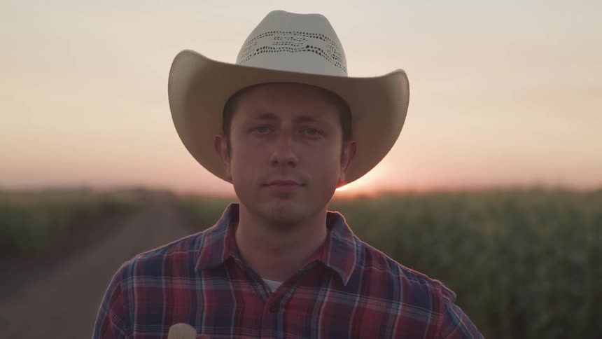 A man wearing a cowboy hat stands beside a rural road on a farm at sunset