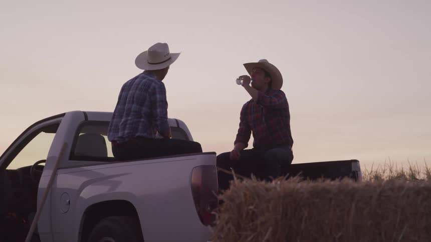 Two men wearing cowboy hats sit on the back of a pickup truck and on hay bales drinking beverages with a field of crops in the background at sunset