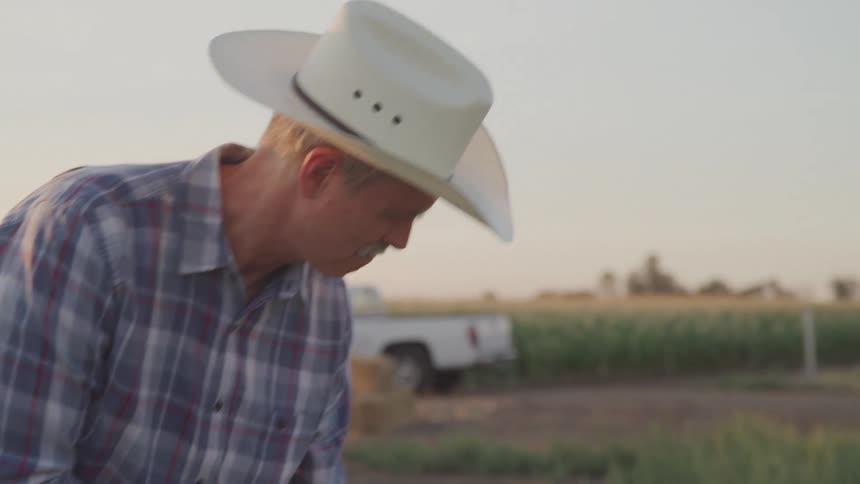 A middleaged man in a cowboy hat and plaid shirt works on a farm at sunset