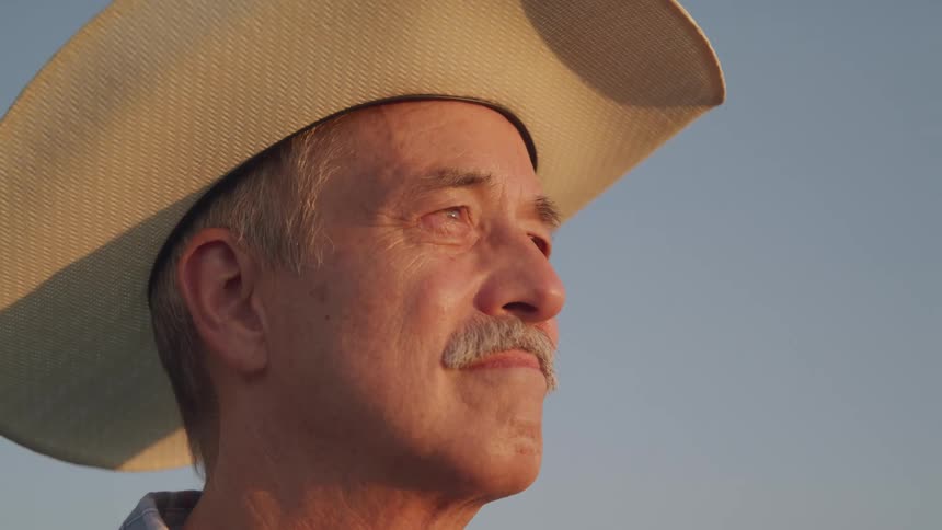 A closeup shot shows a man with a mustache wearing a lightcolored cowboy hat gazing towards the upper right with the lower sun casting warm light on his face against a clear blue sky