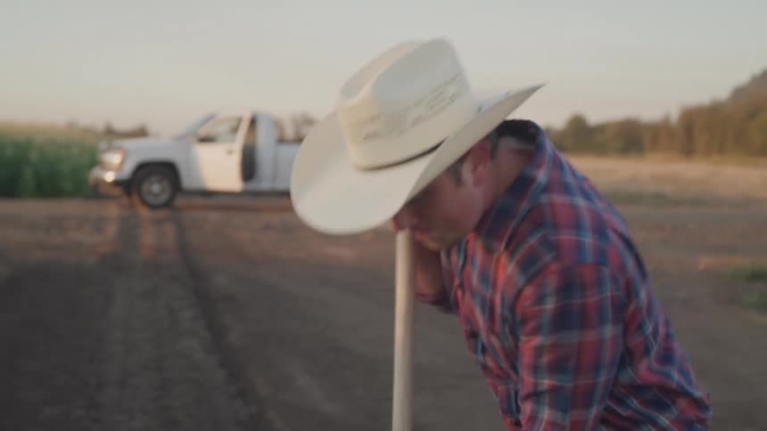 A young man in a cowboy hat and plaid shirt diligently shovels soil on a farm with his pickup truck and hay bales visible in the background