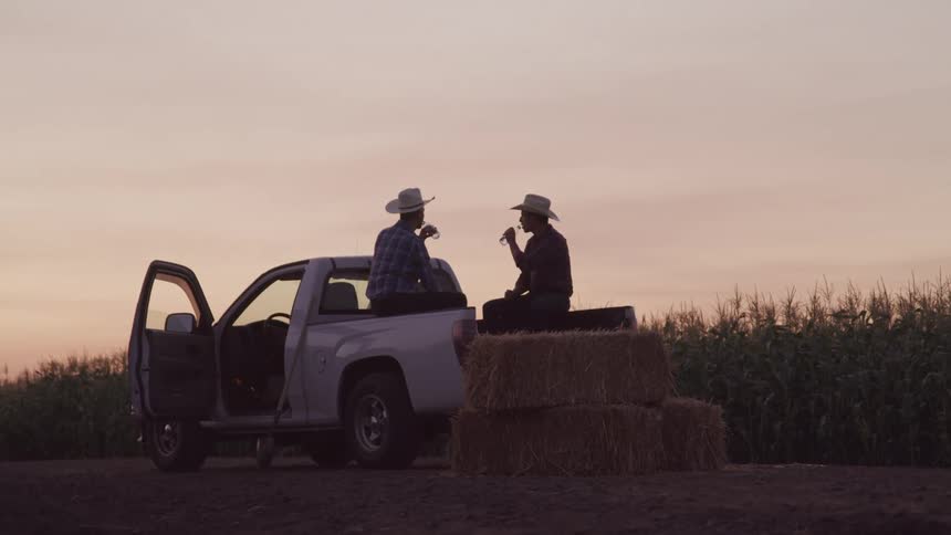 Two men wearing cowboy hats sit on the back of a pickup truck and on hay bales drinking beverages with a field of crops in the background at sunset
