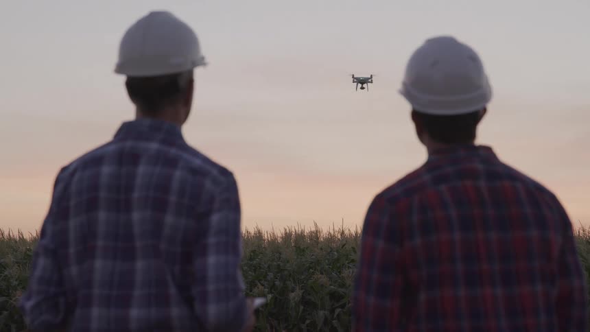 Two men in hard hats monitor a flying drone over a field at sunset