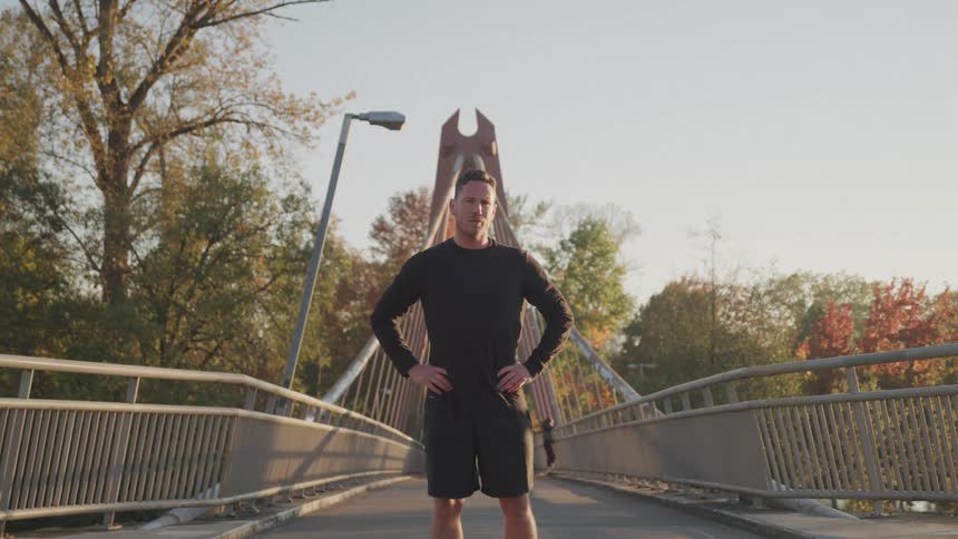 A man in workout clothes standing on a bridge at midday with both hands on his waist after exercising