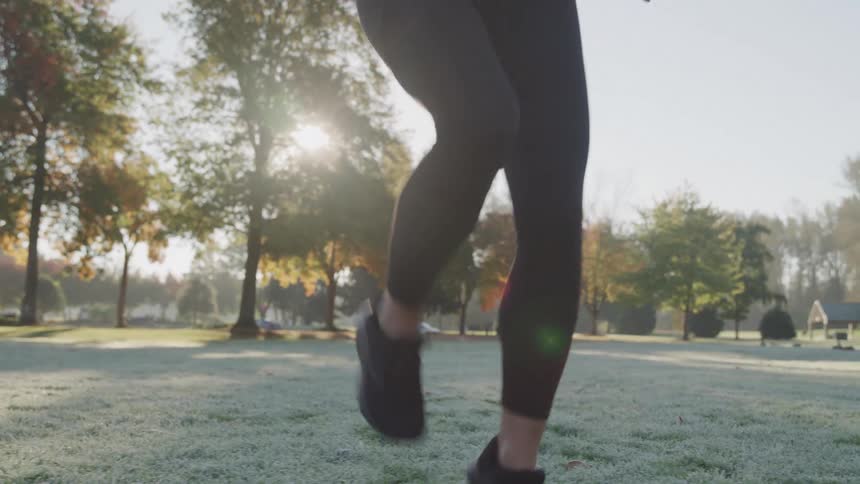 The legs of a woman in black athletic pants and shoes running over orange cones on a grassy field in a park with sunlight filtering through trees