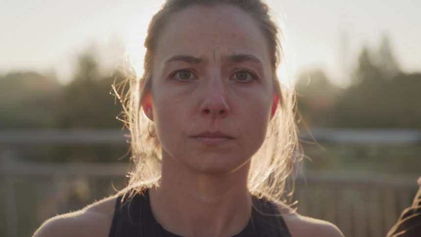 A closeup shot shows a woman with her face partially obscured by the setting sun looking forward