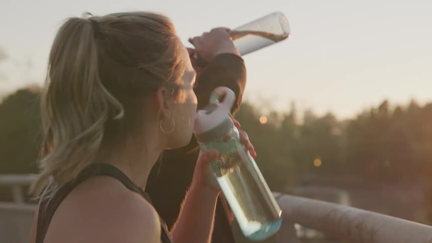 A young man with light brown hair in a ponytail and a man drink from water bottles after a workout on a bridge at sunset