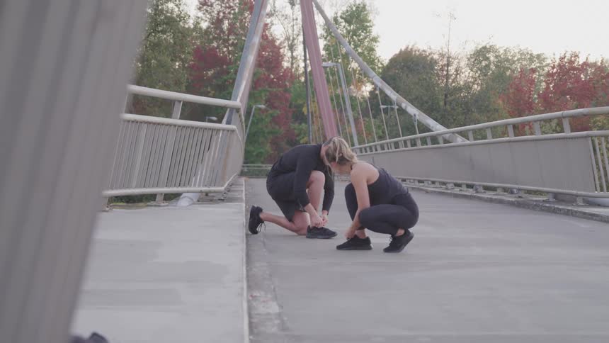 A young man and woman in workout attire tie their shoelaces atop a pedestrian bridge at midday then proceed to stretch together in preparation for exercise