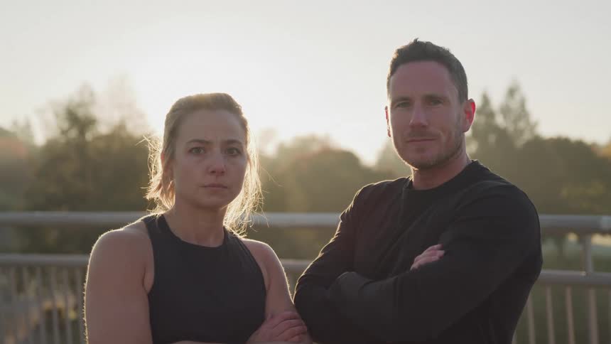 A young man and woman stand sidebyside with their arms crossed looking forward against a sunset backdrop that casts a warm glow over a bridge