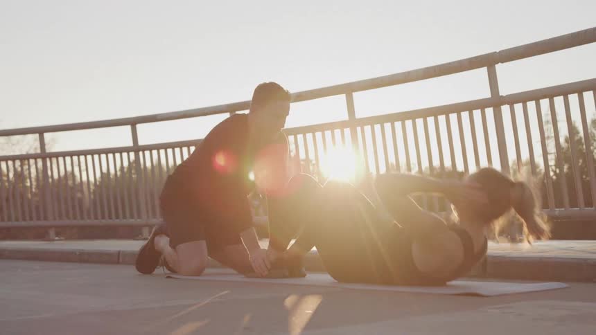 A man helps a woman in a tank topperform crunches on a grey mat on a bridge at sunset