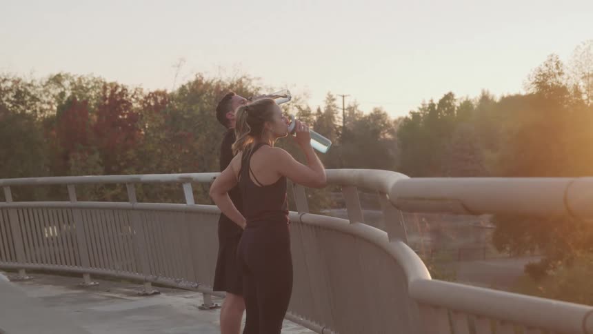 A man and a woman standing by the handrail of a pedestrian bridge after a workout at sunset with the woman sipping water from a water bottle