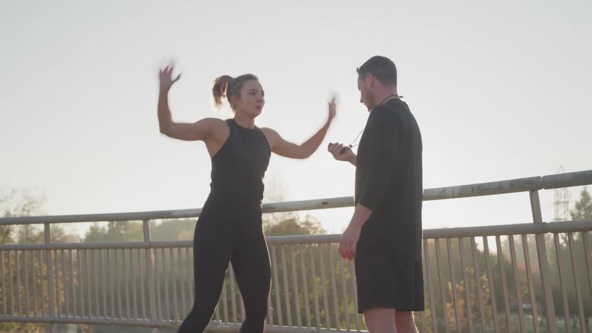 Male white trainer motivates a young female as she performs jumping jacks on a bridge during daytime