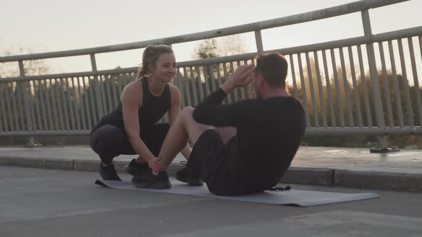 A woman in a black tank top helps a man in a black longsleeved shirt perform crunches on a grey mat on a bridge at sunset