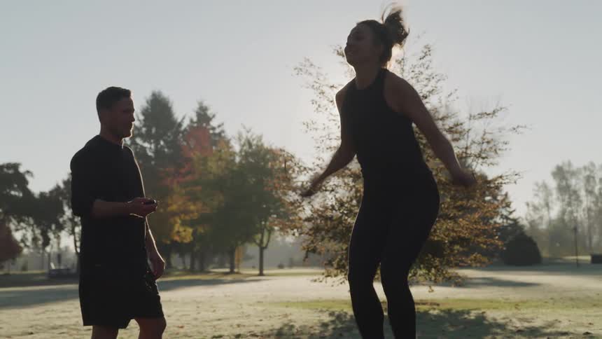 Silhouette of a woman jumping rope outdoors at a park while being timed by her trainer