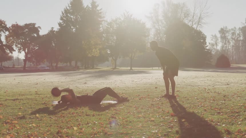 A woman performs crunches on a white mat tossing a black medicine ball to a man standing nearby in a sunlit grassy park with trees