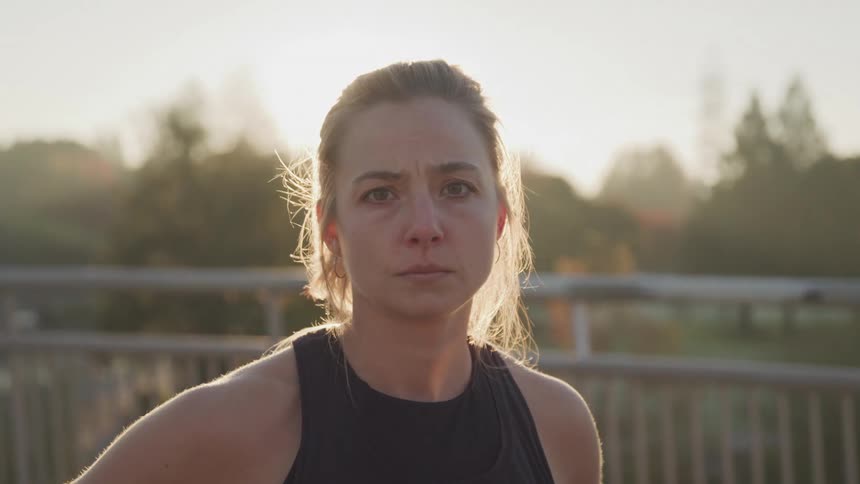 A closeup shot shows a woman with her face partially obscured by the setting sun looking forward with trees and a bridge in the background