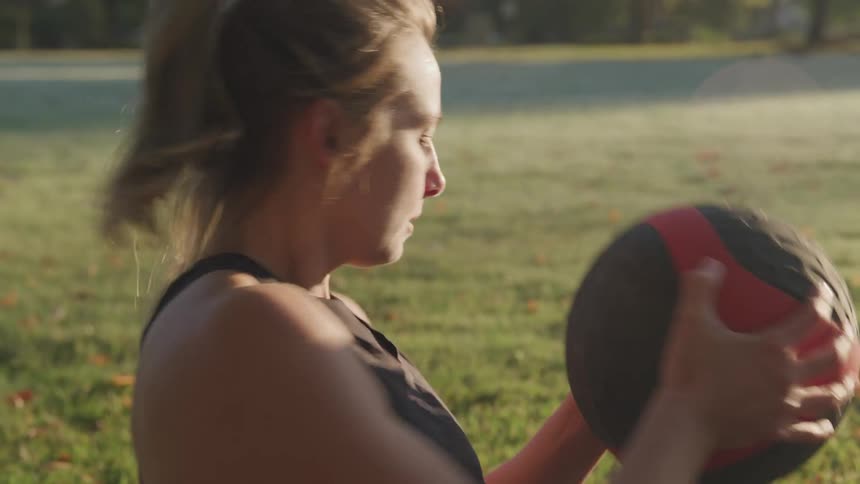 A woman in a black tank top is engaged in a workout holding a black and red medicine ball in her hands with a sunlit grassy field and trees in the background