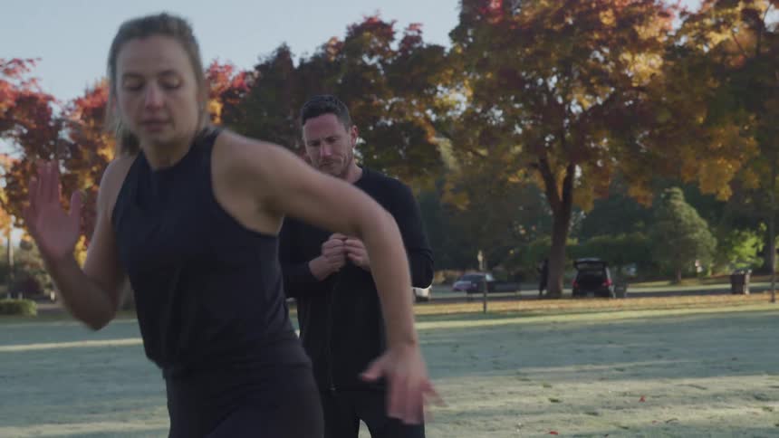A woman running sideways back and forth in a public park at midday with a man on the side cheering and encouraging her during her workout