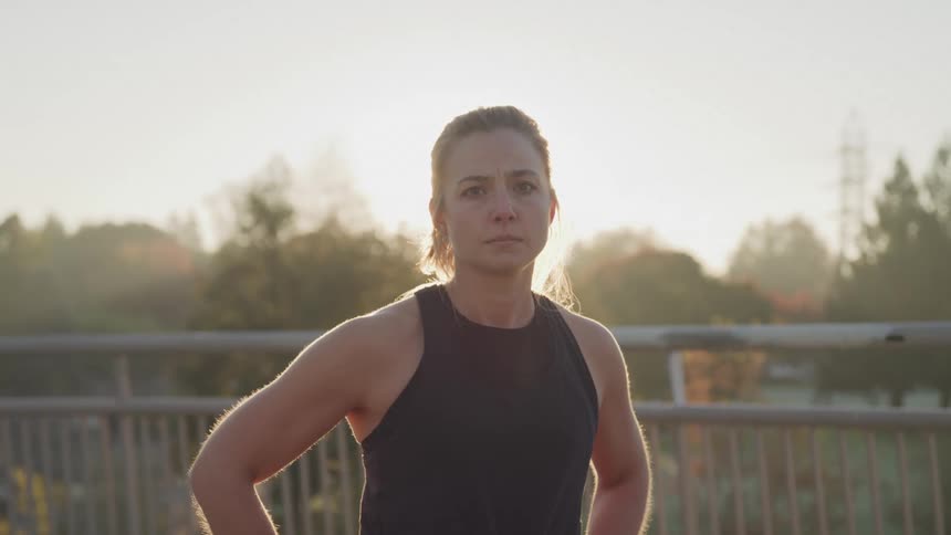 A young woman standing on a bridge at midday wearing a black workout top with her hands on her waist