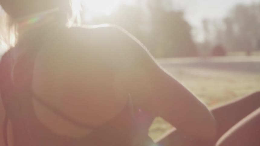 A woman in a black tank top is engaged in a workout holding a black and red medicine ball in her hands with a sunlit grassy field and trees in the background