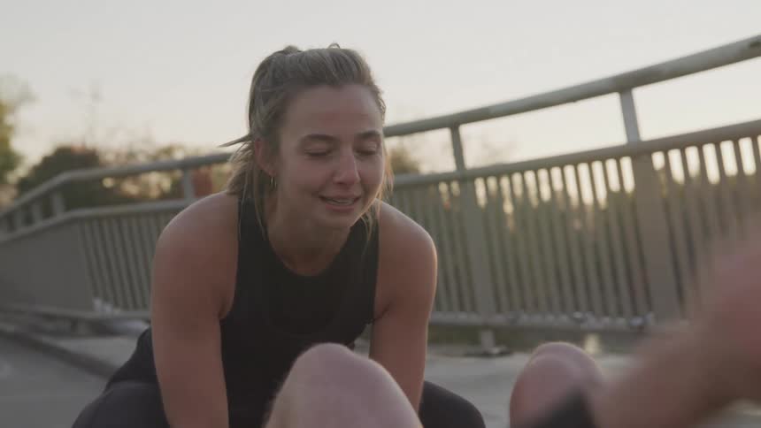 Woman helps a man do sit ups while on a bridge at sunset