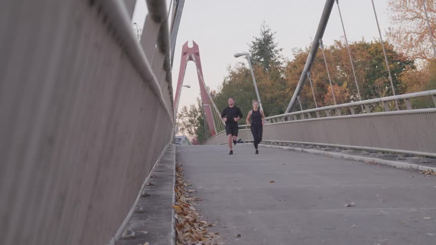 A young couple wearing black tops and black pants running across a bridge at midday