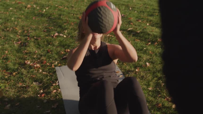 Young woman exercises happily with a trainer by throwing and catching a ball while performing situps on a field during the day