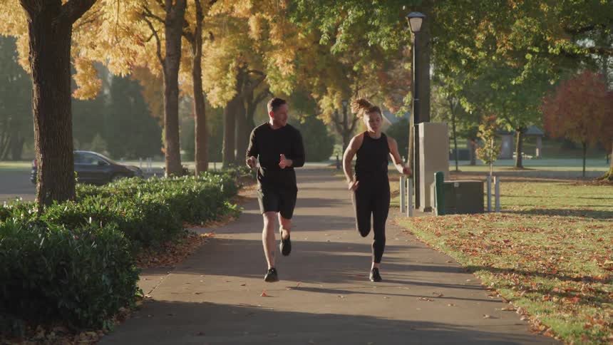Male trainer runs alongside and motivates a young woman as she runs down a pathway during the day